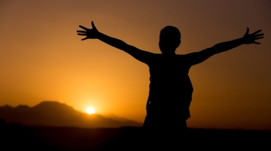 Silhouette of young backpacker person standing with raised arms feeling happy in colorful picturesque landscape, sun rising or setting in mountains on horizon